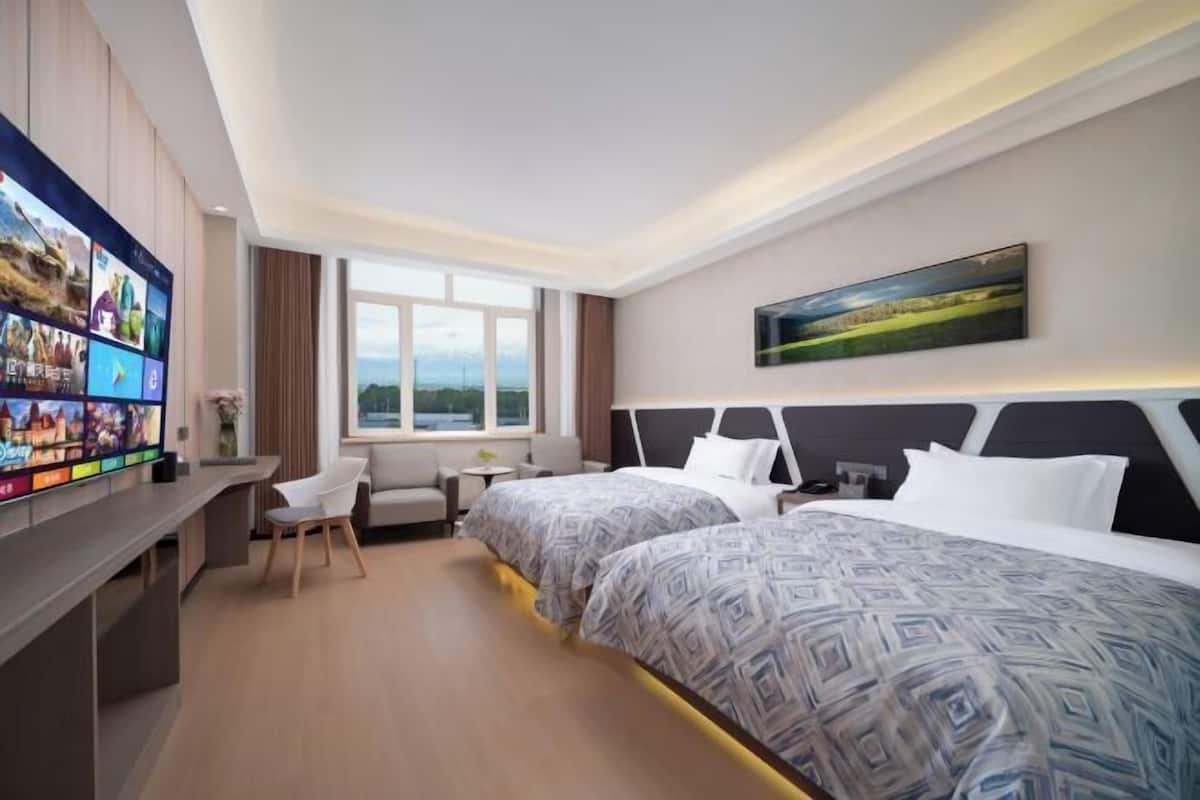 A hotel room with a large TV, two beds, a desk, a chair, a small table, and a window with a view.