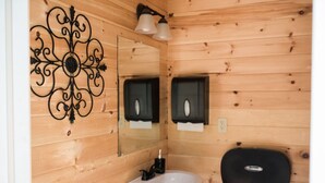 Ein Badezimmer mit Holzwandverkleidung, einem weißen Waschbecken, einem dekorativen Metallwandbild, zwei schwarzen Seifenspendern und einem Spiegel.