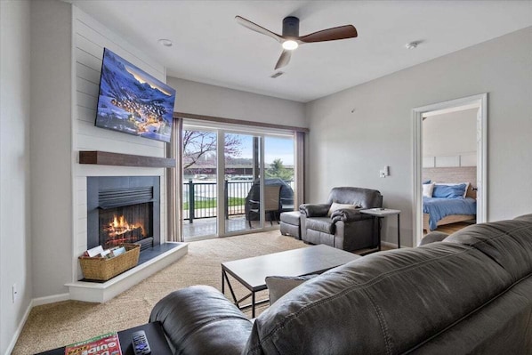 A living room with a fireplace, a leather sofa, a coffee table, a TV mounted above the fireplace, and a ceiling fan.