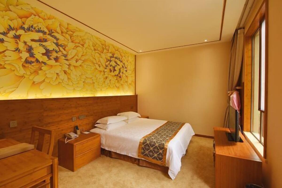 Une chambre d’hôtel avec une grande fresque florale sur le mur, une tête de lit en bois, un lit avec du linge de maison blanc, une table de chevet avec une lampe et une fenêtre avec des rideaux.