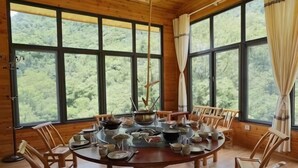 Uma sala de jantar de madeira com uma mesa redonda posta para seis pessoas, rodeada por seis cadeiras, janelas amplas com cortinas e vista para uma floresta.