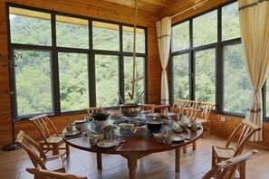 Un comedor de madera con una mesa redonda preparada para seis comensales, rodeada por seis sillas, ventanas amplias con cortinas y vistas a un bosque.