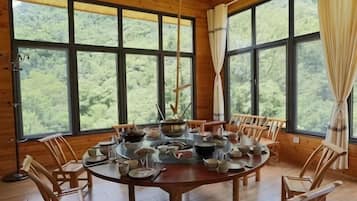 Comedor de madera con una gran mesa redonda dispuesta para una comida, rodeada de sillas, ventanas amplias y cortinas.
