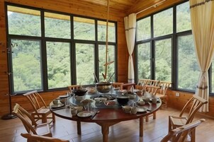 Comedor de madera con una gran mesa redonda dispuesta para una comida, rodeada de sillas, ventanas amplias y cortinas.