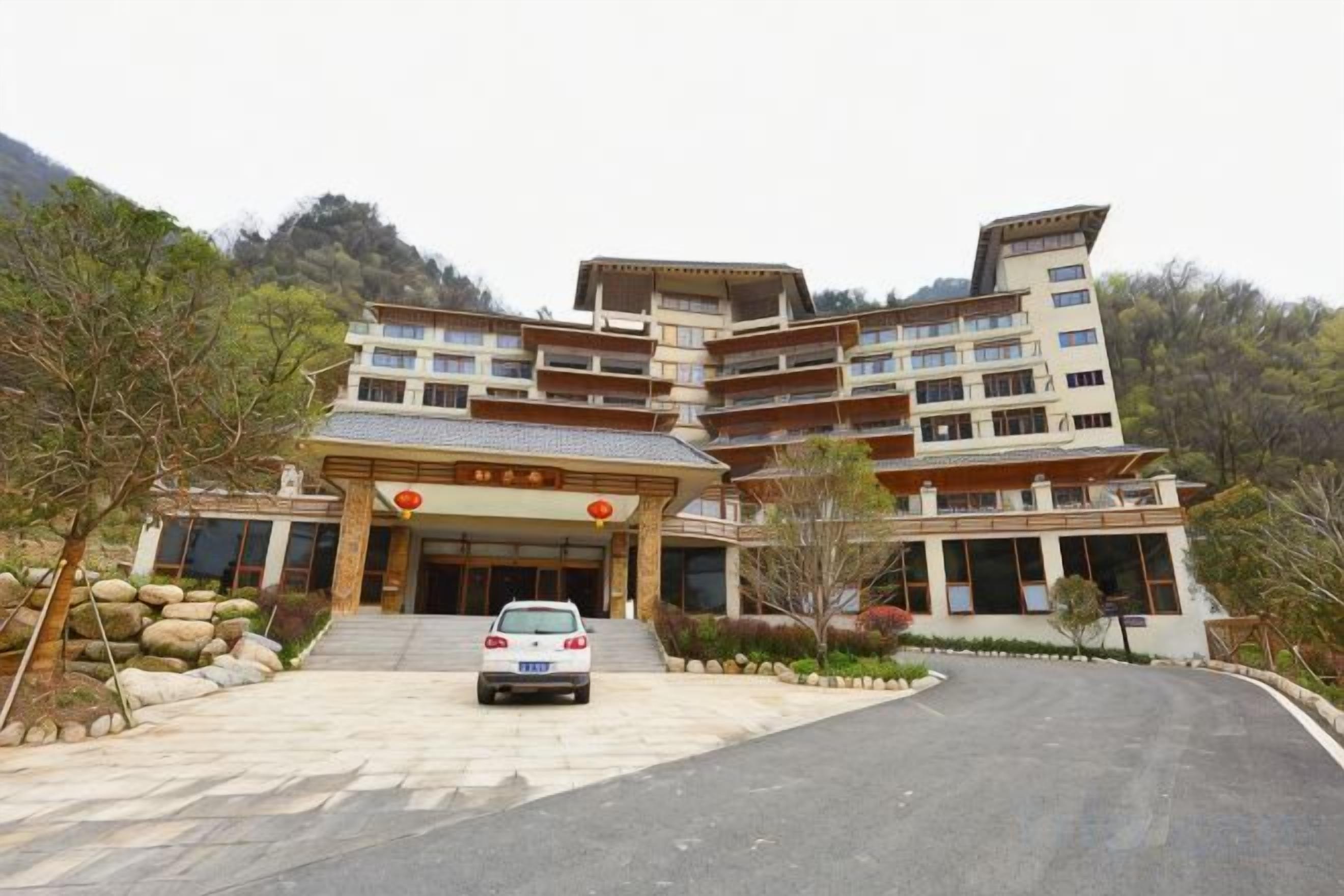 A multi-story hotel with a traditional architectural style, featuring a central entrance with steps, a parking area in front, and a mountainous backdrop.