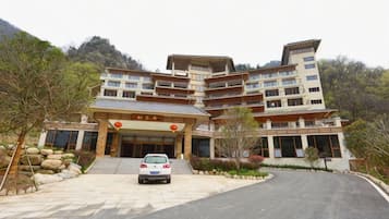 A multi-story hotel with a traditional architectural style, featuring a central entrance with steps, a parking area in front, and a mountainous backdrop.