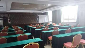 Un salón de conferencias con filas de mesas y sillas, una pantalla de proyector y un amplio ventanal.
