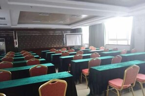 Uma sala de conferências com fileiras de mesas e cadeiras, uma tela de projetor e uma janela ampla.