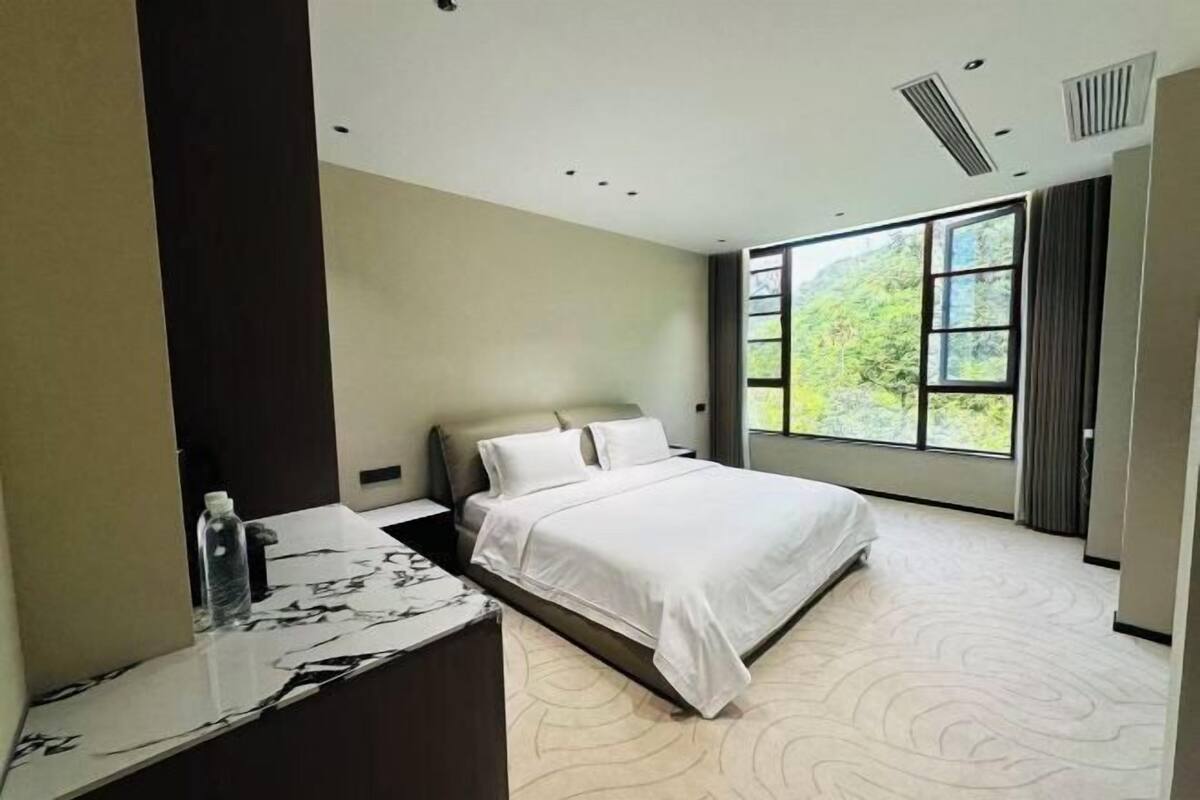 Une chambre d’hôtel moderne avec un grand lit, une table de chevet en marbre, une grande fenêtre donnant sur un paysage verdoyant et une bouche d’aération au plafond.