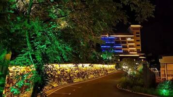 Una carretera curva que conduce a un edificio de varios pisos, rodeado de vegetación e iluminado con luz artificial.