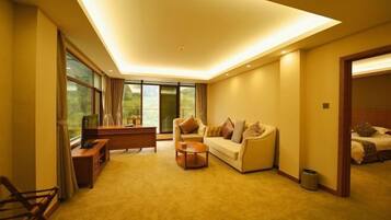 A modern hotel room with a sofa, a coffee table, a TV, and a large window offering a view of the outdoors.