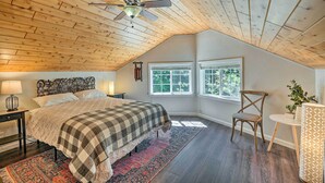 Un dormitorio con techo inclinado, una cama con colcha a cuadros, una silla de madera, una mesita con lámpara y una ventana con vistas a zonas verdes.