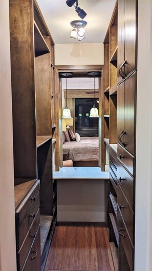 A narrow walk-through closet with wooden shelving and drawers, leading to a bedroom with a bed and nightstands.