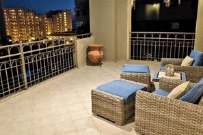 Un balcón con mobiliario de mimbre, un jarrón grande y vistas de edificios de la ciudad por la noche.