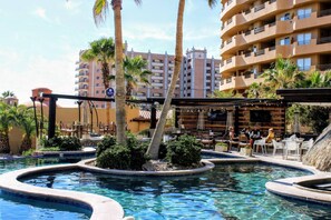 Zona de piscina en un resort con palmeras, un bar y un edificio al fondo.
