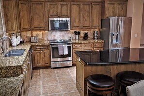 Una cocina con gabinetes de madera, electrodomésticos de acero inoxidable y encimeras de granito, además de una isla central con taburetes.