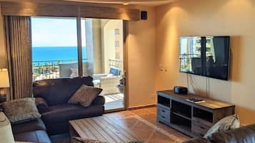 A living room with a sofa, coffee table, and TV. It has a balcony with a view of the sea.