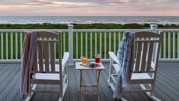Two rocking chairs on a wooden deck with a table between them, overlooking a beach with a railing and a view of the ocean.
