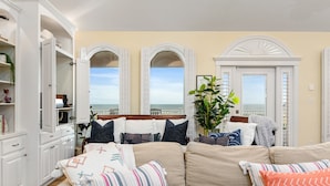 A bedroom with a beige sofa, a large window with a view of the sea, and white cabinetry.