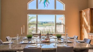 Un coin repas avec une grande table dressée pour six personnes, une vue sur la campagne par une grande fenêtre et un buffet en bois sur le côté droit.