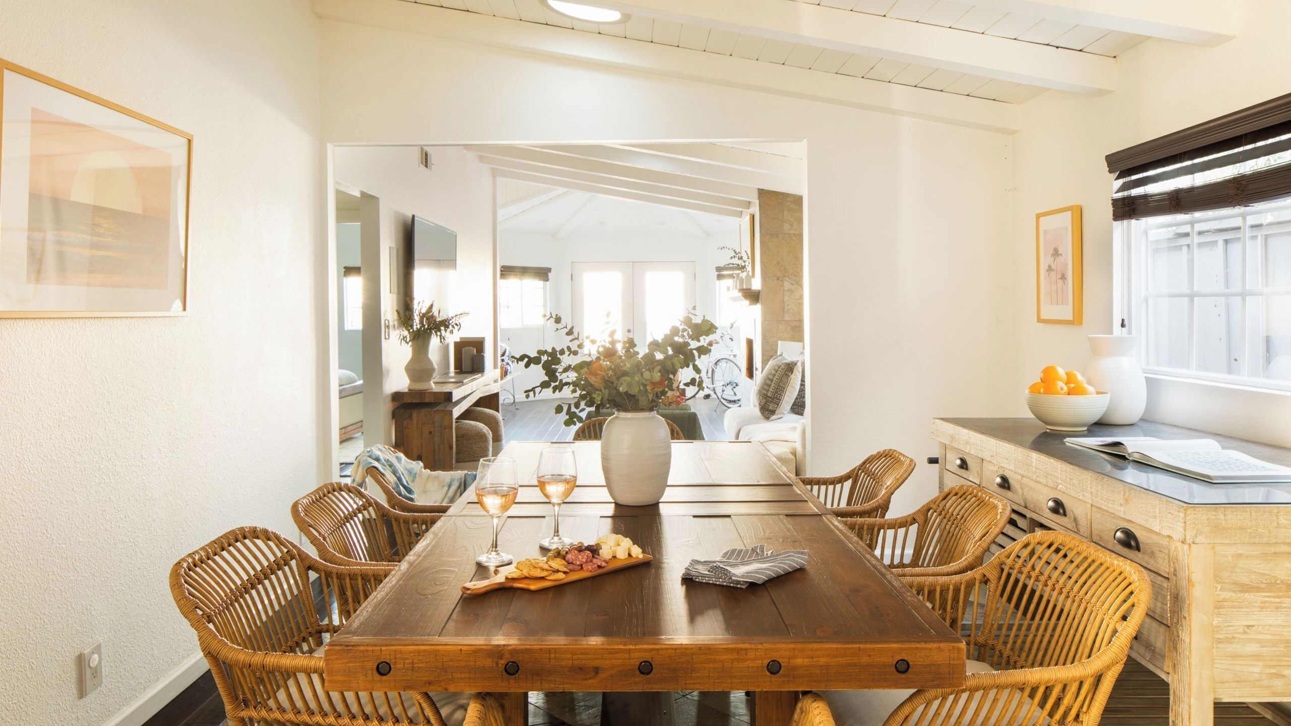 A dining area with a wooden table, wicker chairs, a vase with flowers, and a bowl of fruit.