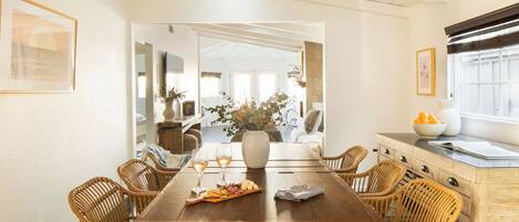 A dining area with a wooden table, wicker chairs, a vase with flowers, and a bowl of fruit.