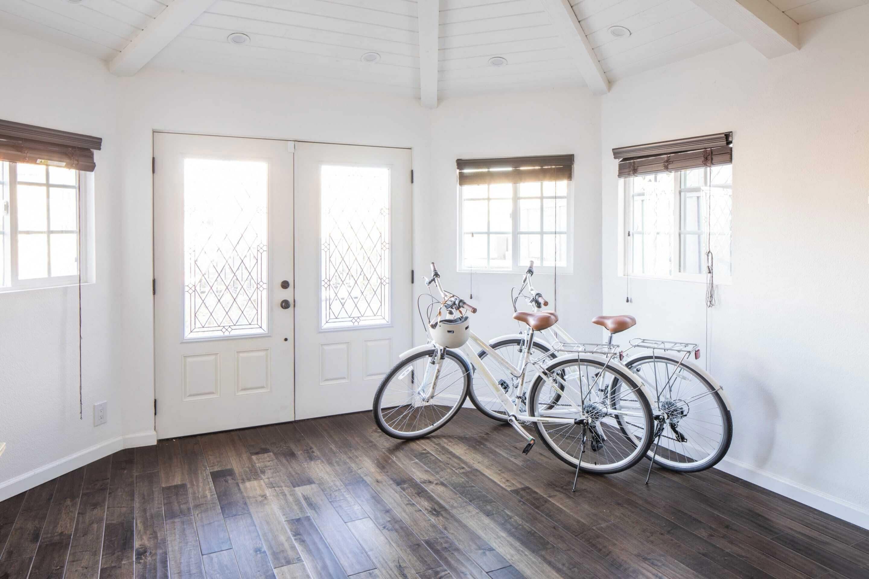 Een ruime kamer met donkere houten vloer, witte muren en een hoge plafond. Er zijn twee witte deuren met ruitpanelen en twee ramen met bruine luiken. Tegen de muur staan twee fietsen geparkeerd.