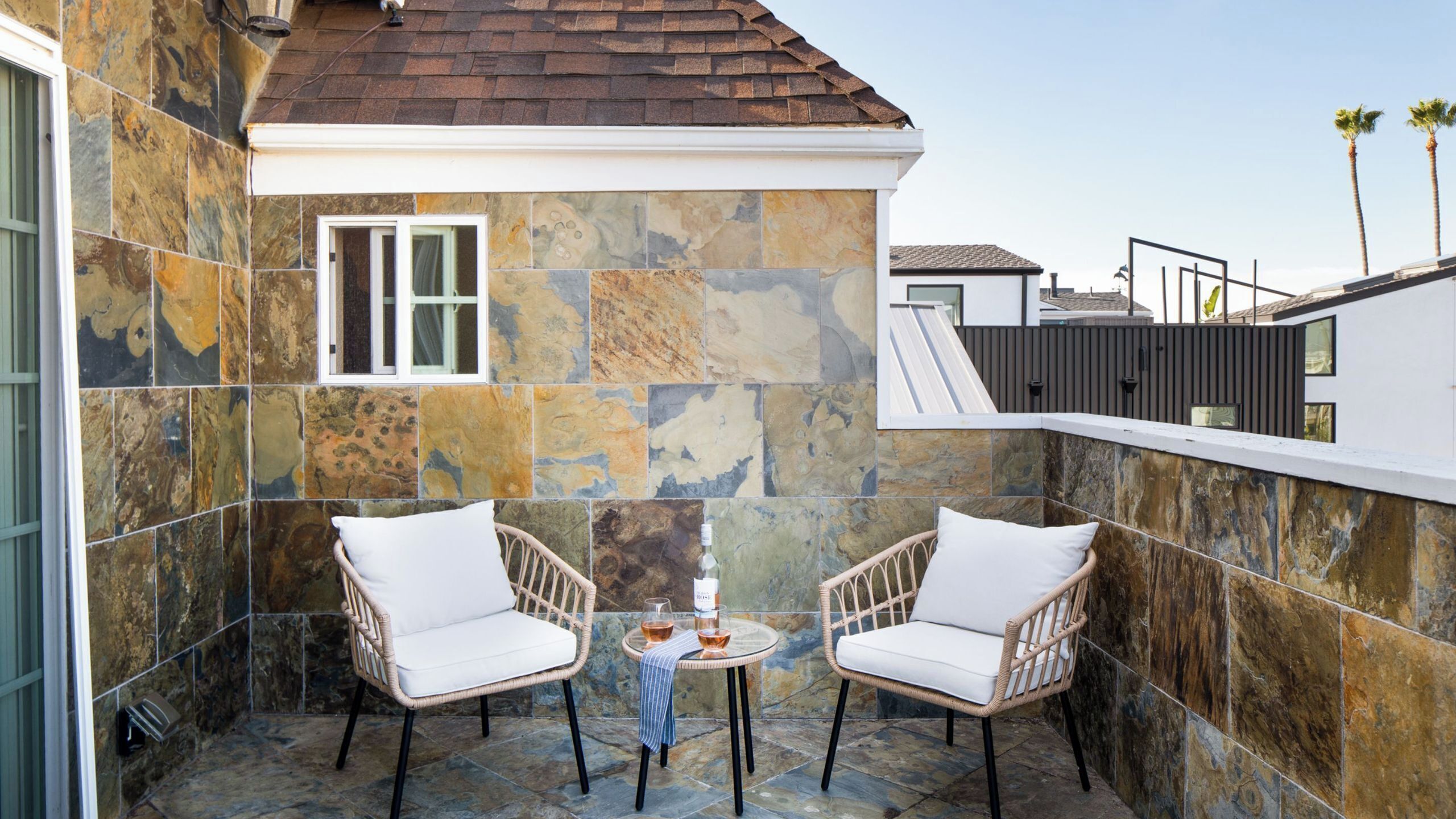 A balcony with stone tiles, two wicker chairs, a small round table with a bottle and glass, and a window.