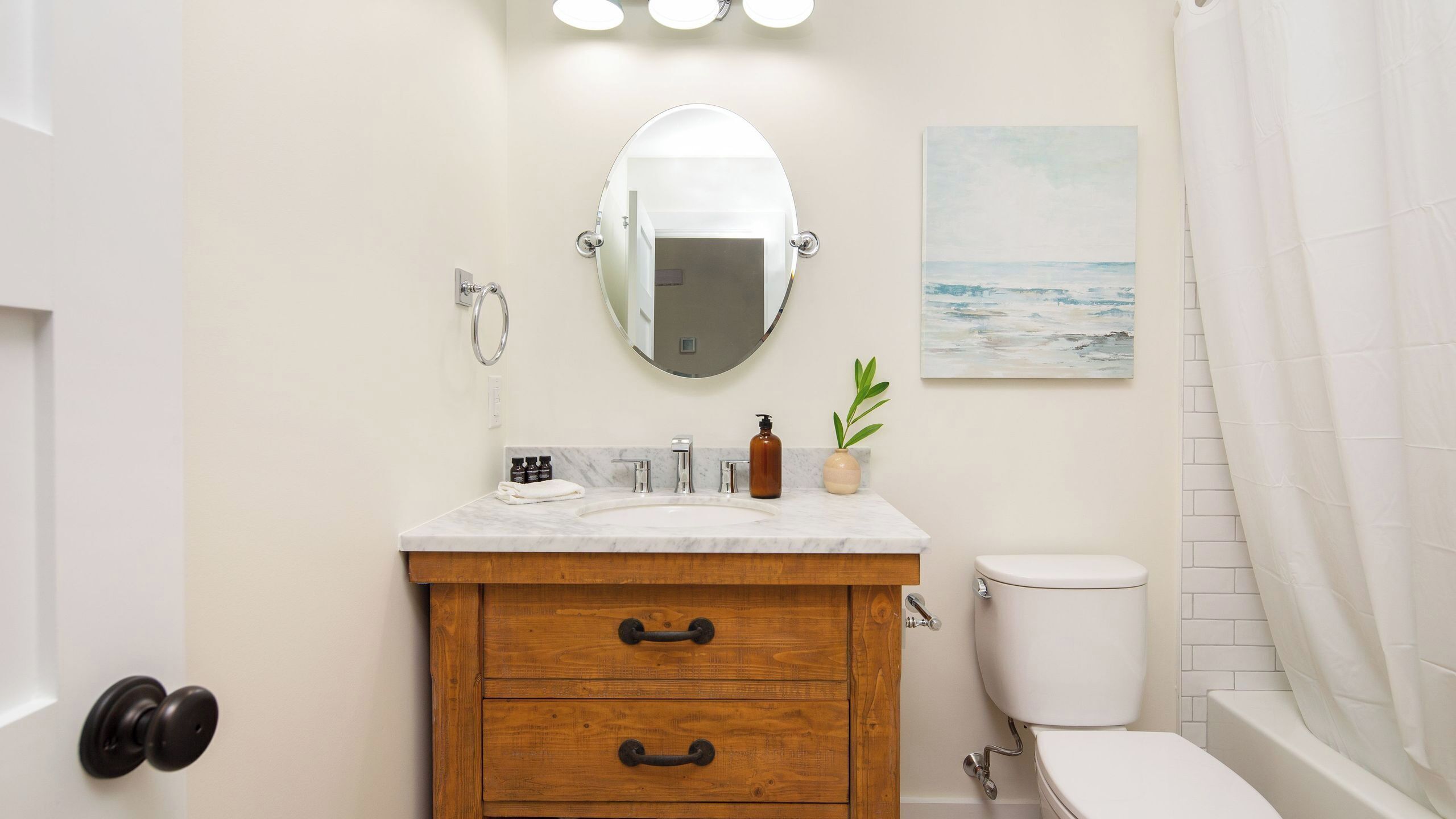 Une salle de bain avec un meuble-lavabo en bois, un miroir rond, un lavabo blanc, des toilettes et un tableau représentant une plage.
