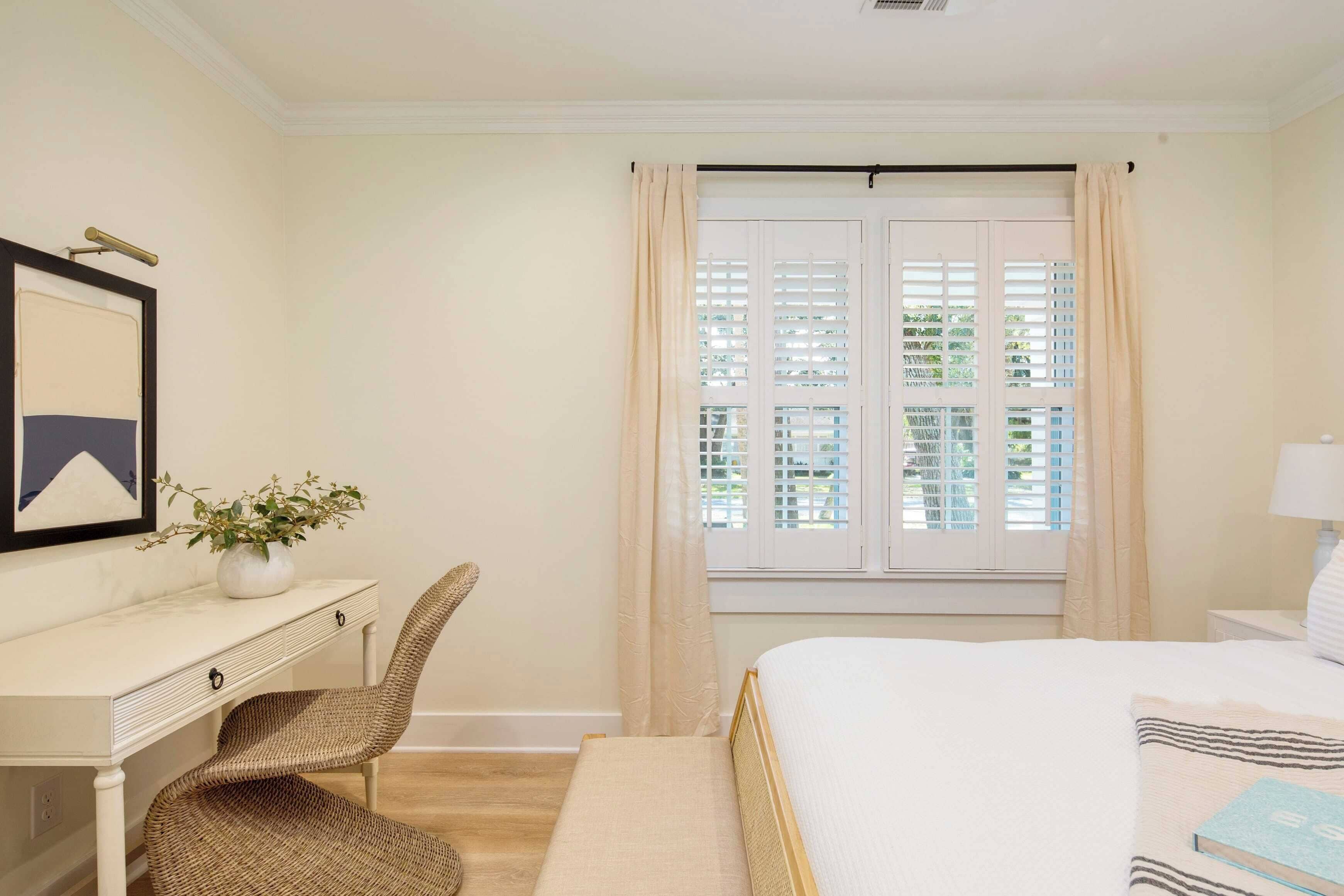 A bedroom with a bed, a desk with a chair, a window with curtains, and a framed picture on the wall.
