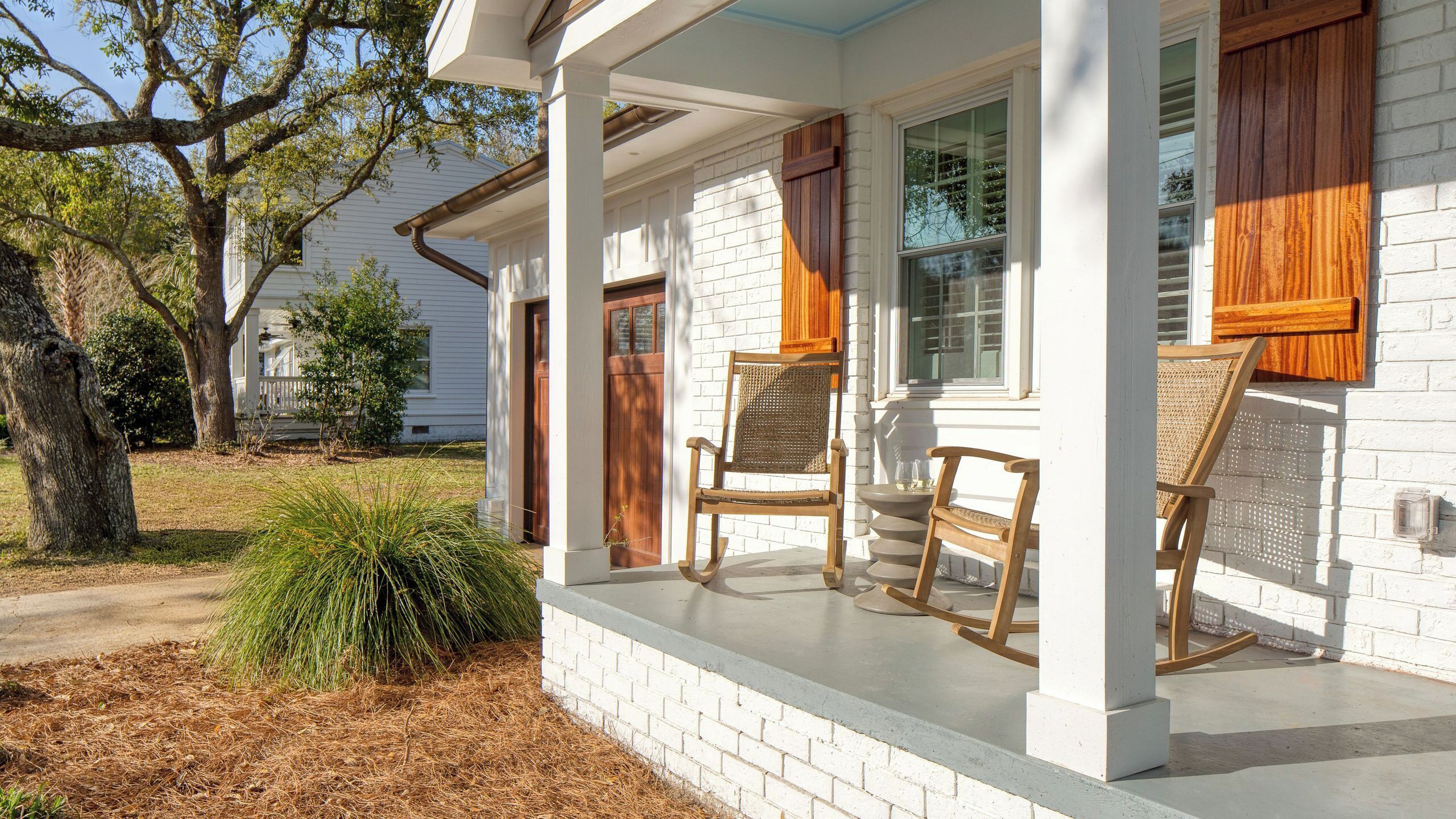 A porch with two rocking chairs, a brick wall, and a white house with wooden shutters.
