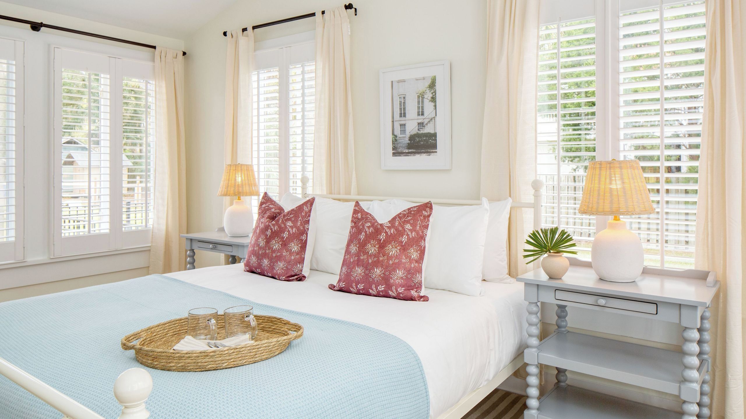 Une chambre à coucher comprenant un lit, deux tables de chevet, une commode, une lampe, un tableau encadré et une fenêtre avec des stores.