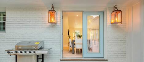 Un patio con una parrilla de acero inoxidable, un banco de madera y un ventanal que da a un espacio interior con una mesa y sillas.