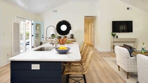 A modern kitchen with a central island, a dining area with wicker chairs, and a flat-screen TV mounted on the wall. - Sawyer by Avantstay Stunning Isle Of Palms Home w/ Pool! (Isle of Palms)