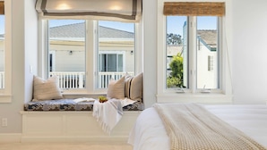 A bedroom with a large window, built-in bench with cushions, and a bed with a white comforter. - Crystal Cove by Avantstay Gorgeous & Charming Newport Home! (Newport Beach)