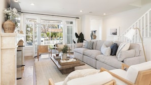 A spacious living room with a large grey sofa, a coffee table, and a fireplace. There is a staircase with a white railing and a framed picture on the wall. - Crystal Cove by Avantstay Gorgeous & Charming Newport Home! (Newport Beach)