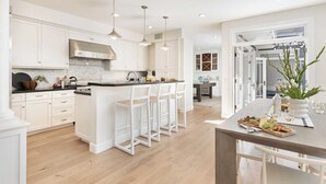 A modern kitchen with white cabinetry, a central island with bar stools, and a dining area with a table set for two. - Crystal Cove by Avantstay Gorgeous & Charming Newport Home! (Newport Beach)