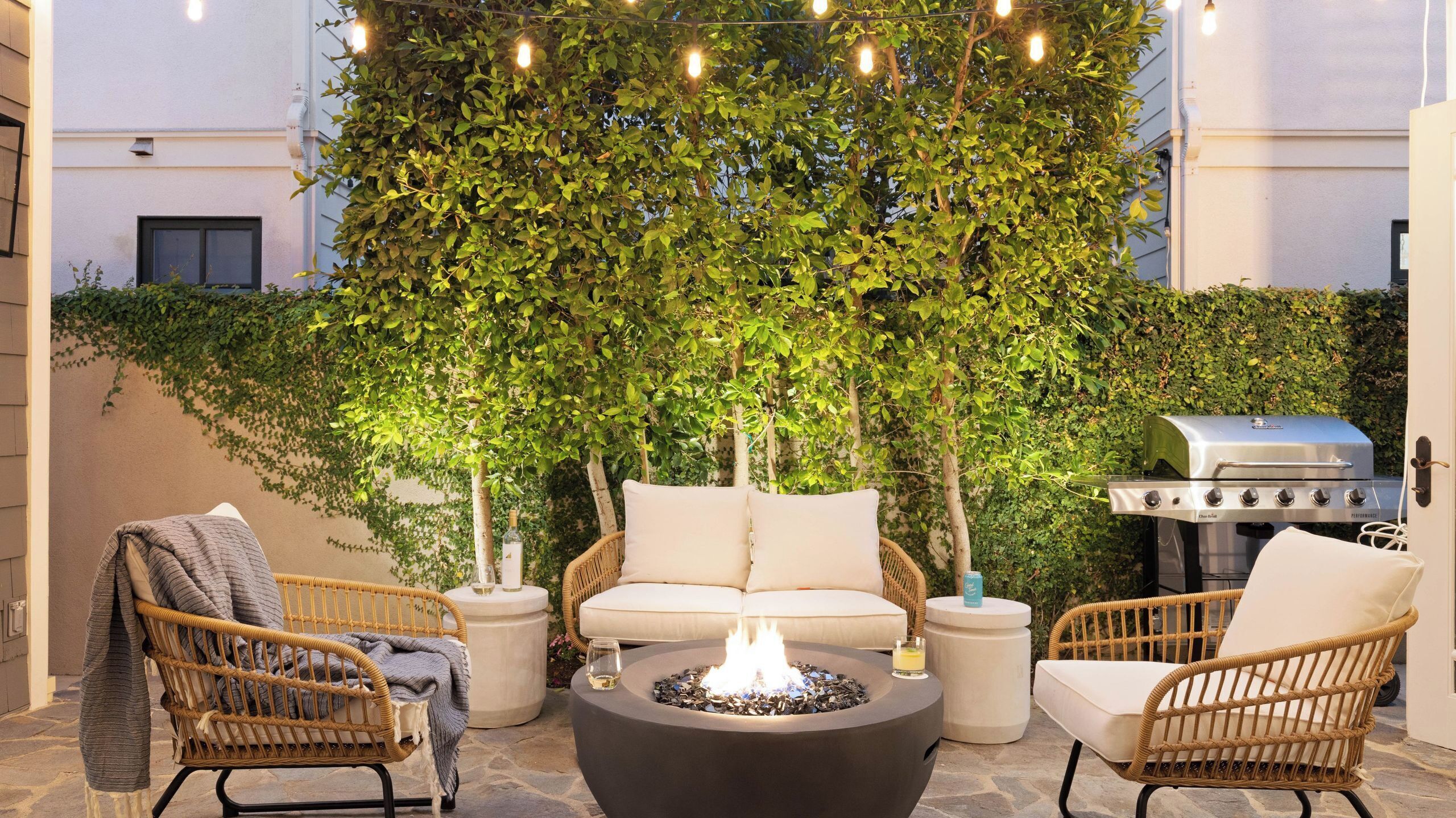 An outdoor seating area with wicker chairs, a round table with a fire pit, and a grill.