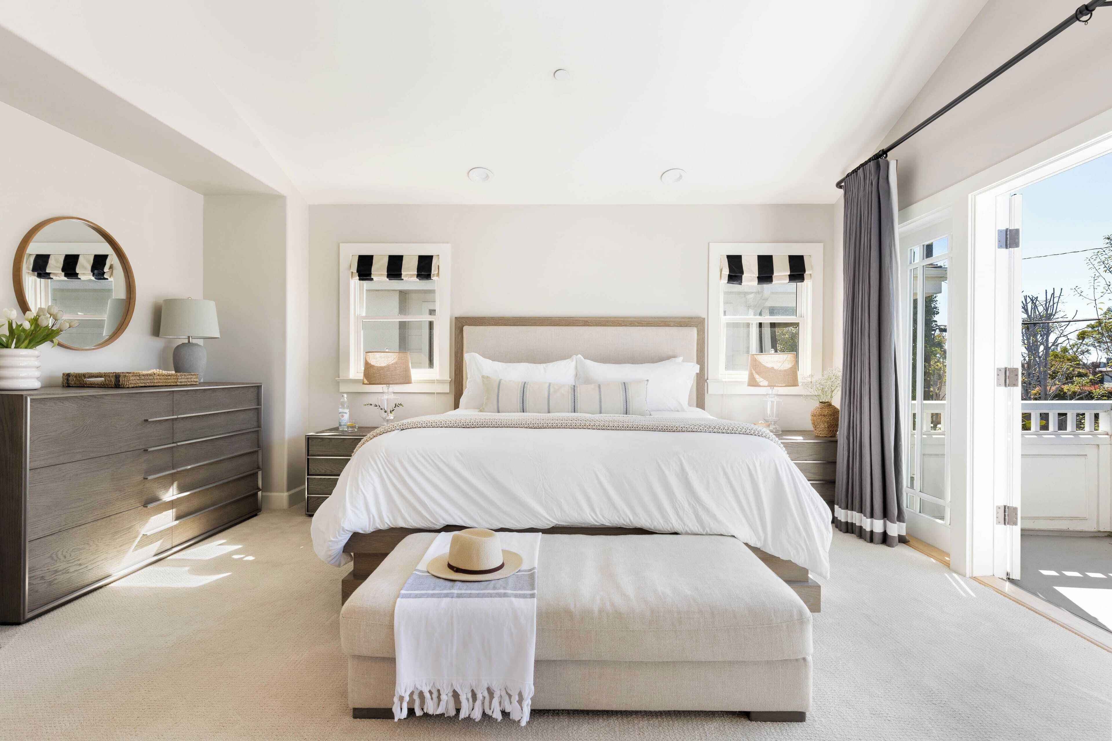 A bedroom with a large bed, bedside tables, a dresser, a mirror, and a view of the outdoors.