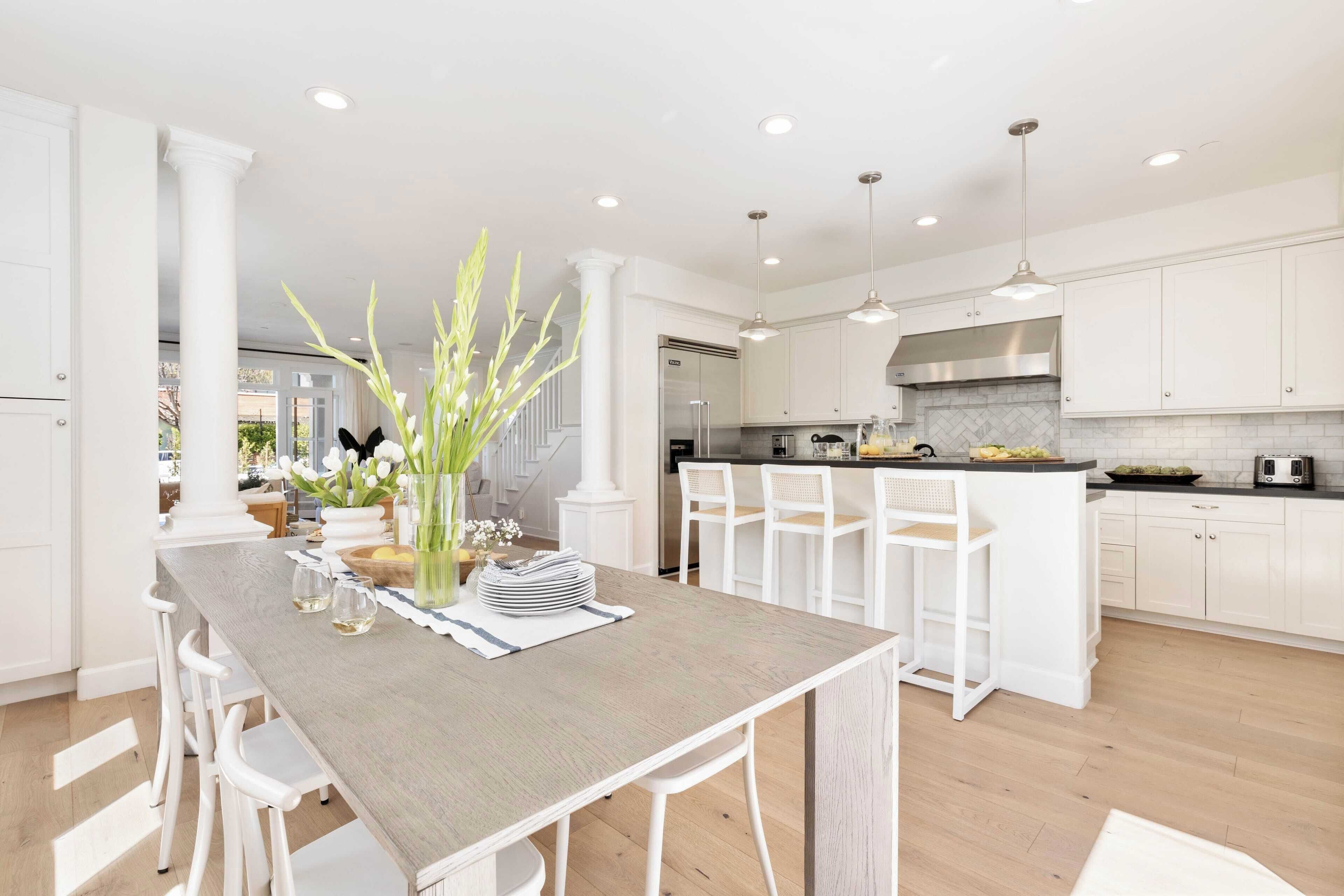 A modern kitchen with white cabinetry, a central island with bar stools, and a dining area with a long table and white chairs.