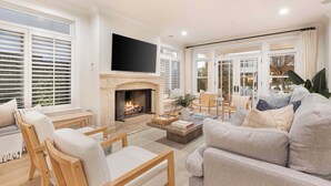A living room with a fireplace, a television, and a large window with blinds. - Crystal Cove by Avantstay Gorgeous & Charming Newport Home! (Newport Beach)