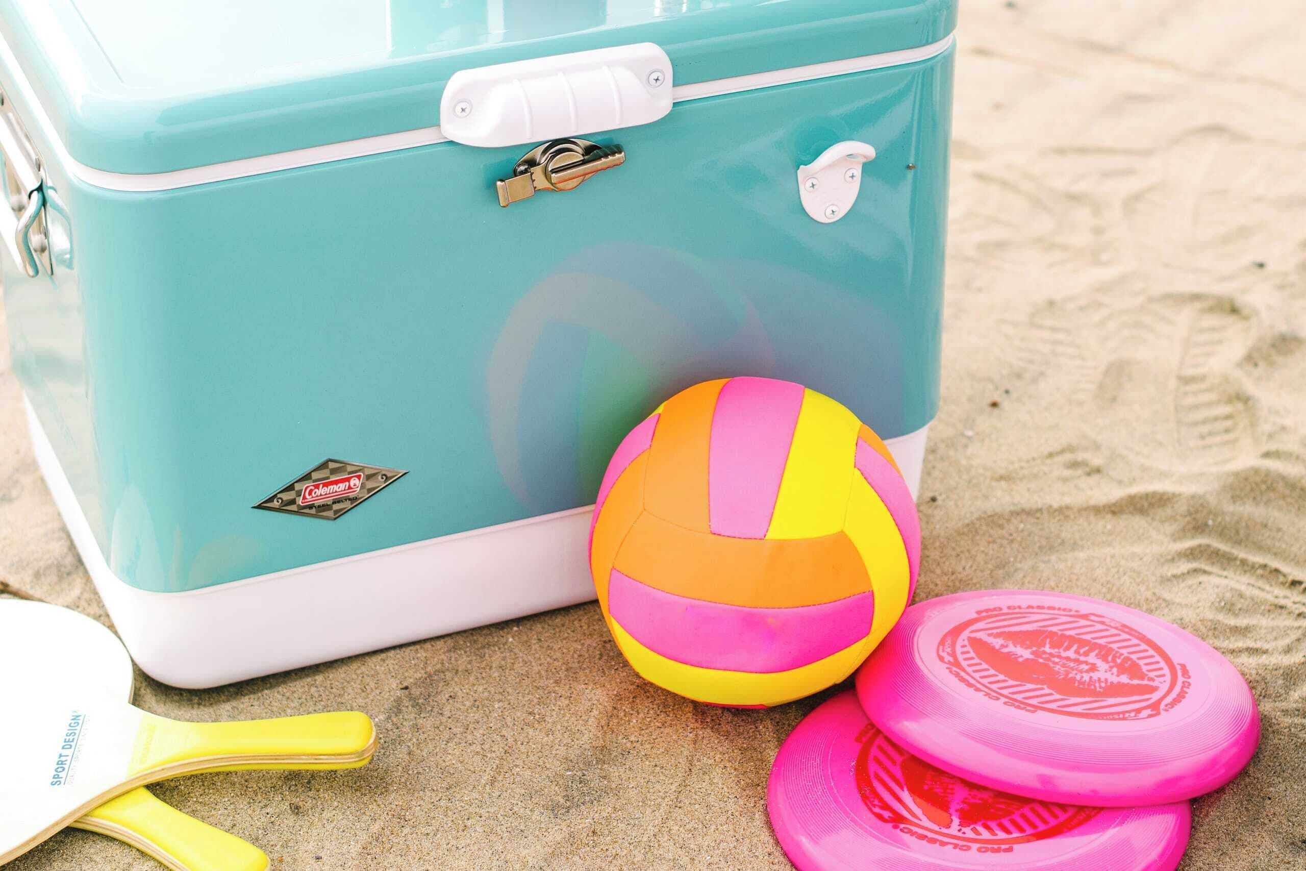 Uma mala azul-turquesa com o logotipo da Coleman, uma bola de praia e dois frisbees cor-de-rosa sobre uma superfície de areia.