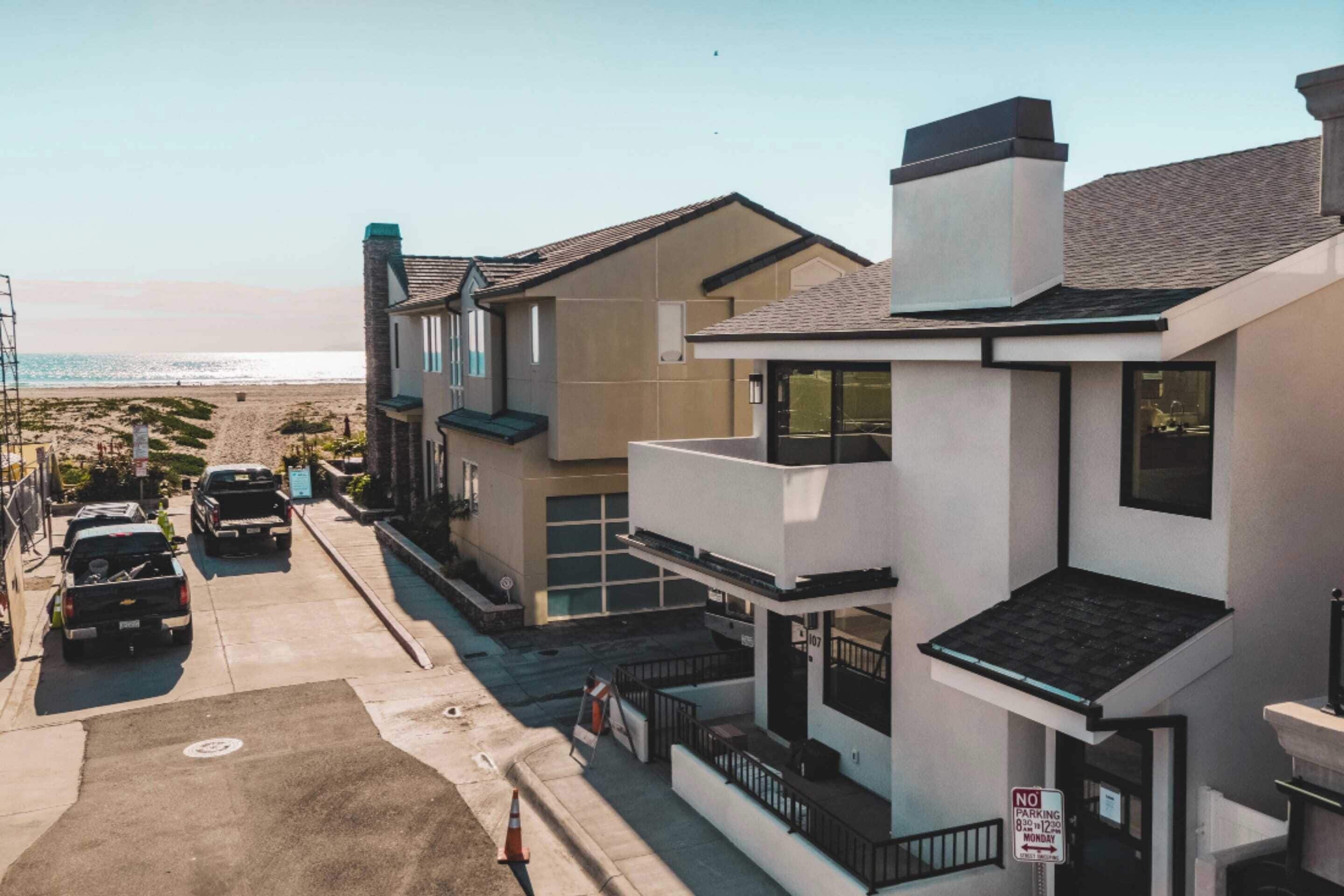 A coastal street with modern houses, a clear view of the beach, and a 'No Parking' sign.