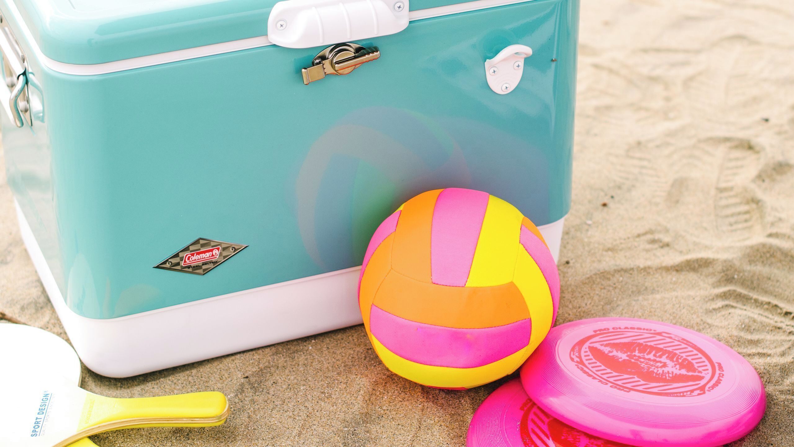 A turquoise suitcase, a beach ball, and a pink frisbee on sandy ground.