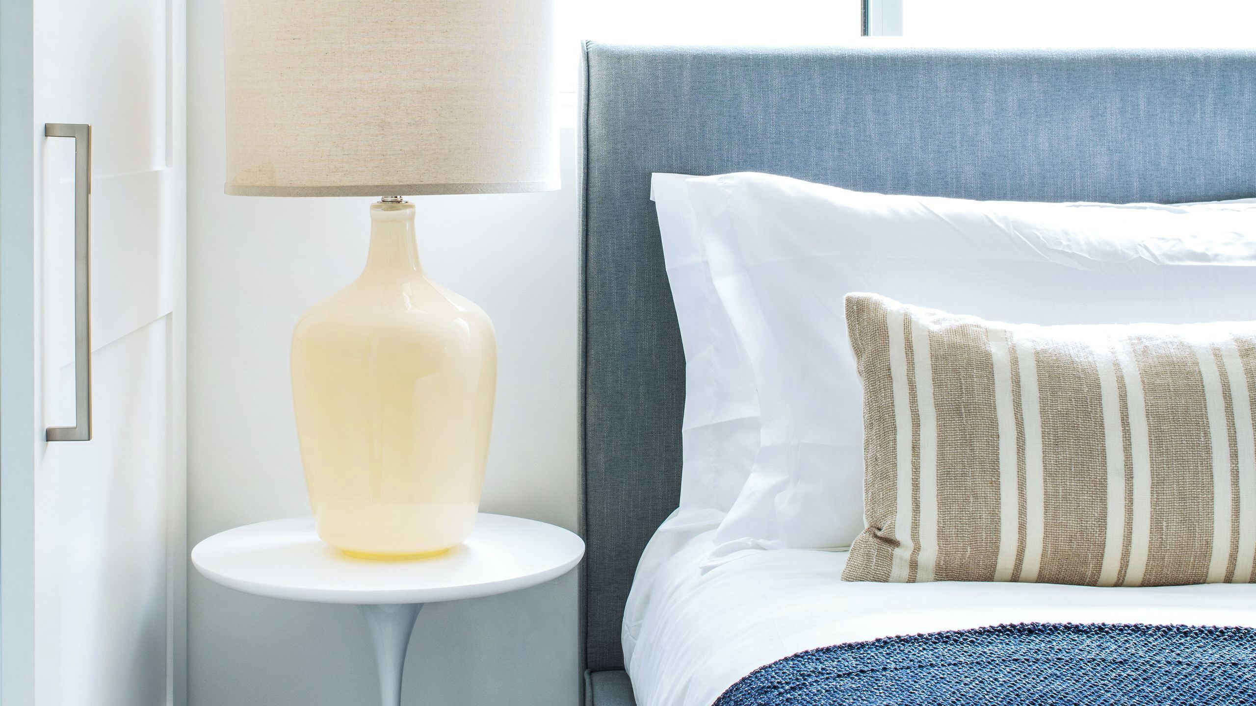 Una cama bien hecha con almohadas blancas y a rayas, un cabecero gris y una mesita de noche pequeña de color blanco con una lámpara.
