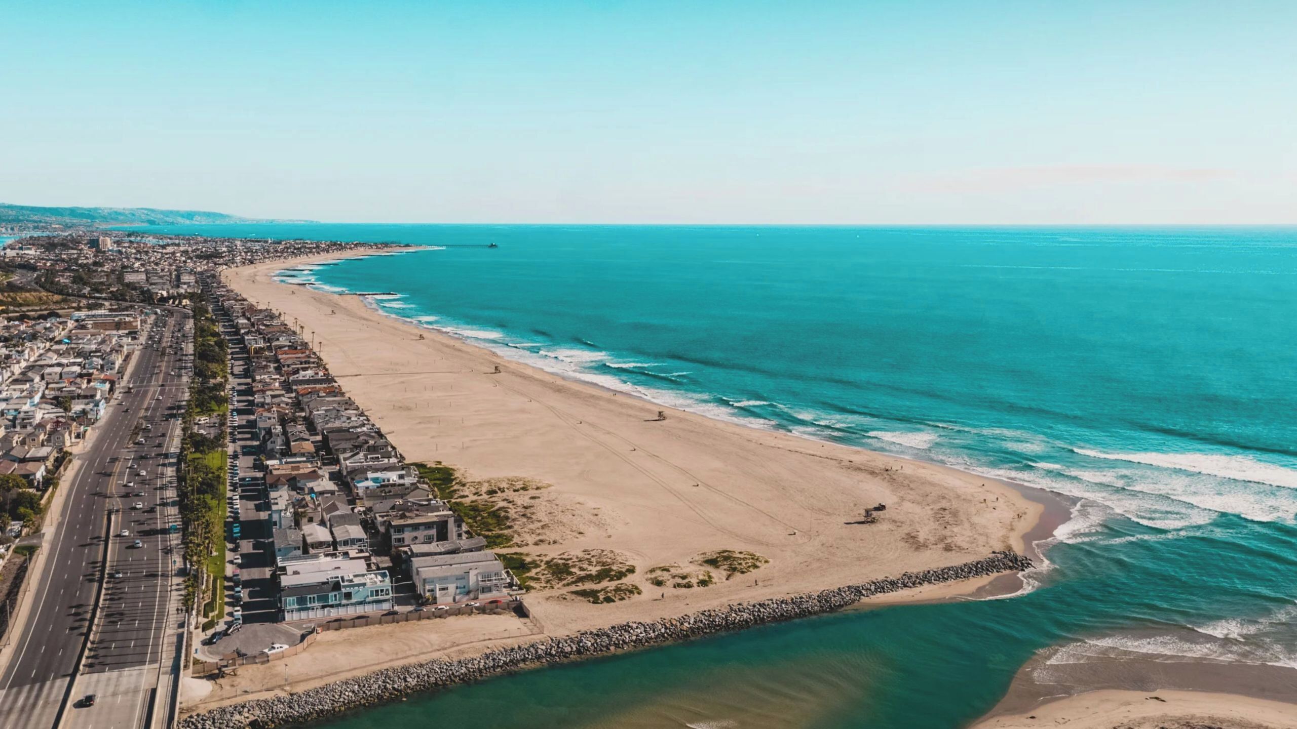 An aerial view of a coastal city with a long beach, a road parallel to the shore, and a rocky breakwater.