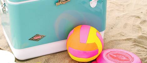 Una maleta color turquesa con el logotipo de Coleman, una pelota de playa y dos frisbees rosas sobre arena.