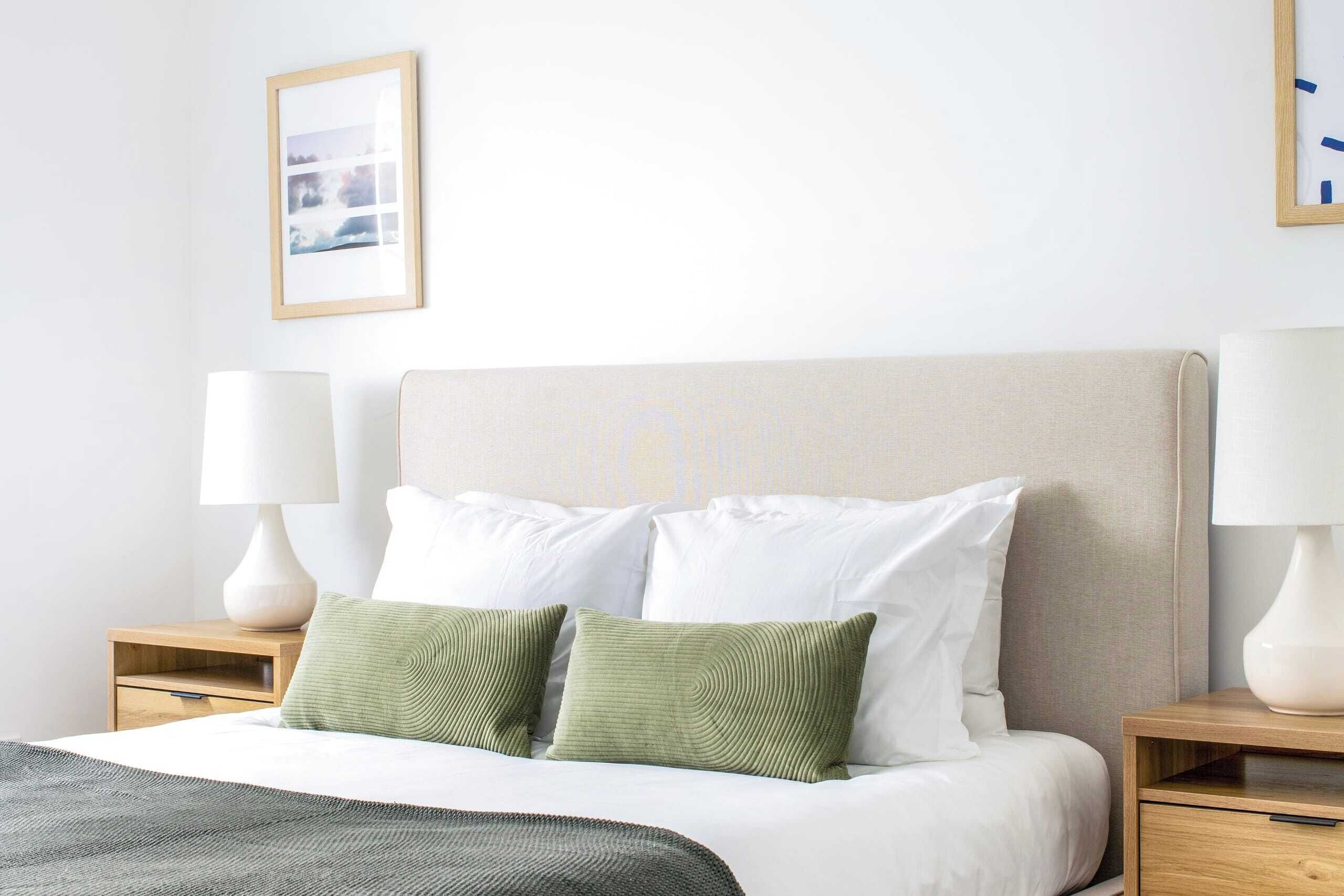 A neatly made bed with white bedding and two green pillows, flanked by wooden nightstands with white lamps.