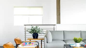 Uma sala de estar moderna com um sofá cinzento, uma mesa de centro de madeira, uma mesinha redonda com um vaso azul e livros, e uma janela com persianas.