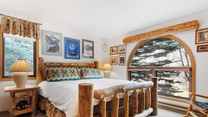 A cozy bedroom with a wooden bed, a large window with a view of trees, and various framed pictures on the wall. - Riverside Condos B101 by Avantstay Condo Close To Downtown, Town Park & Ski Lift #8 (Telluride)